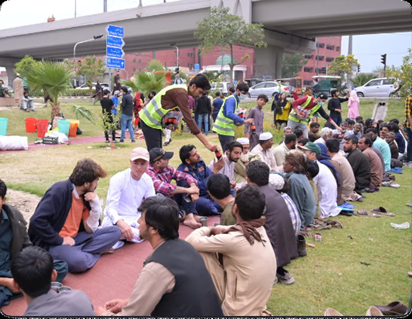 Volunteers in green Rizq vests serving people seated in rows on a grass verge beside a road in Lahore