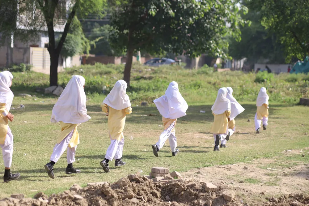 Students in school uniform running across an open field near the school, seen from behind and at a distance